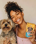 Woman holding a dog and a package of probiotics for dogs supplement in a pouch, smiling at the camera.